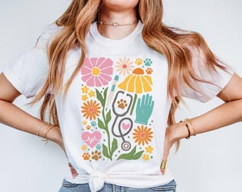 May include: White t-shirt with a colourful floral and medical-themed graphic. The design includes flowers, paw prints, a stethoscope, a thermometer, and a heart rate monitor. The shirt is tied at the waist.