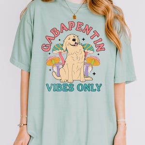 K&ouml;nnte beinhalten: Ein hellgr&uuml;nes T-Shirt mit einem Aufdruck eines Golden Retrievers, der vor drei Pilzen sitzt. Der Text "Gabapentin Vibes Only" ist auf dem T-Shirt aufgedruckt.