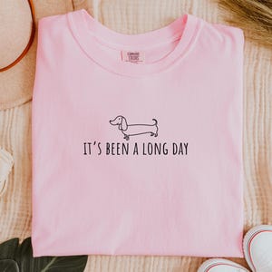 May include: A pale pink t-shirt featuring the phrase "IT'S BEEN A LONG DAY" and a black dachshund outline. The shirt is folded and has a "Comfort Colors" label.