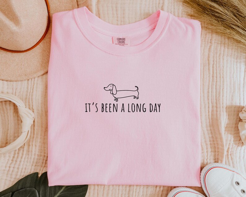 May include: A pale pink t-shirt featuring the phrase "IT'S BEEN A LONG DAY" and a black dachshund outline. The shirt is folded and has a "Comfort Colors" label.