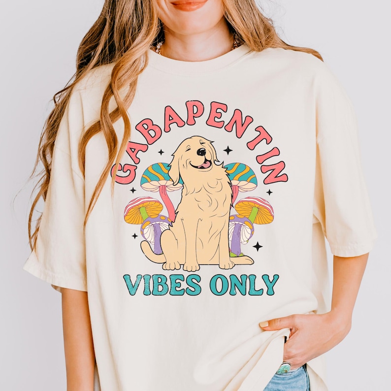 K&ouml;nnte beinhalten: Ein cremefarbenes T-Shirt mit einem Grafikdruck eines Golden Retrievers, der vor bunten Pilzen sitzt. Der Text "Gabapentin Vibes Only" ist in einem Kreis um den Hund gedruckt.