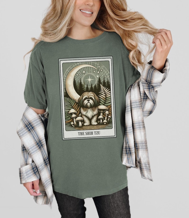 May include: A green t-shirt with a graphic of a Shih Tzu dog in a forest setting with mushrooms and a crescent moon. The text "THE SHIH TZU" is printed below the image.