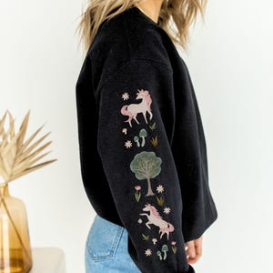 May include: Black sweatshirt with a unicorn, tree, and mushroom design on the sleeve. The design is in pink, green, and white.