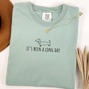 May include: A pale green t-shirt featuring the phrase "It's been a long day" and a dachshund graphic. A gold necklace and a brown hat are also visible. The shirt is made of a soft material.