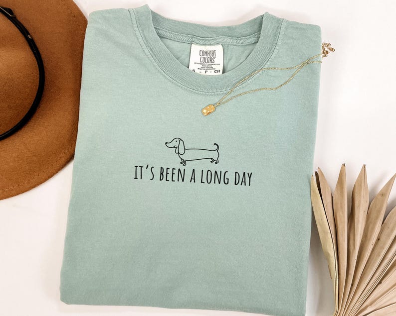 May include: A pale green t-shirt featuring the phrase "It's been a long day" and a dachshund graphic. A gold necklace and a brown hat are also visible. The shirt is made of a soft material.