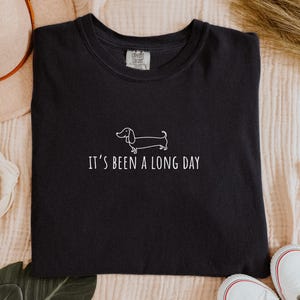 May include: Black t-shirt featuring a white dachshund dog illustration and the phrase "It's been a long day." The shirt has a crew neck.