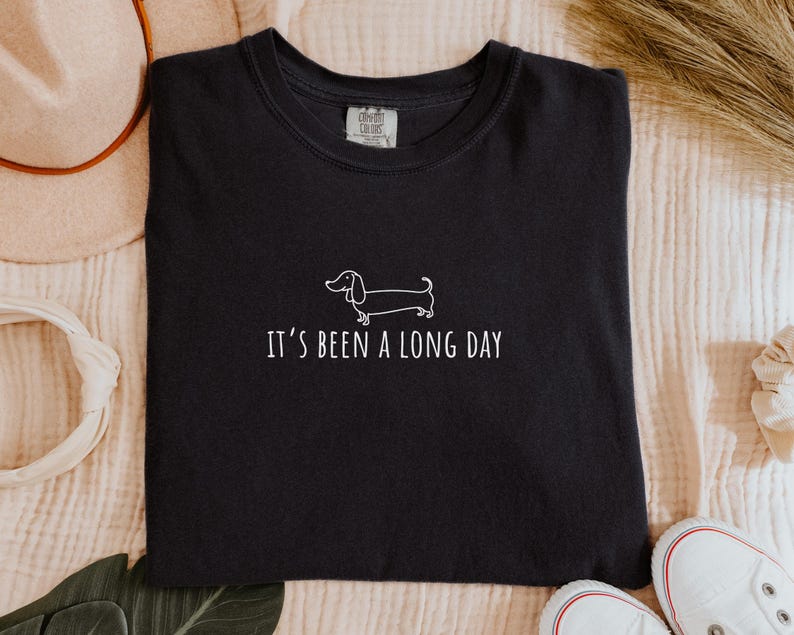 May include: Black t-shirt featuring a white dachshund dog illustration and the phrase "It's been a long day." The shirt has a crew neck.