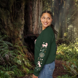 May include: A dark green sweatshirt with long sleeves featuring a pink unicorn and floral design. The sweatshirt is worn by a person standing in a forest setting.