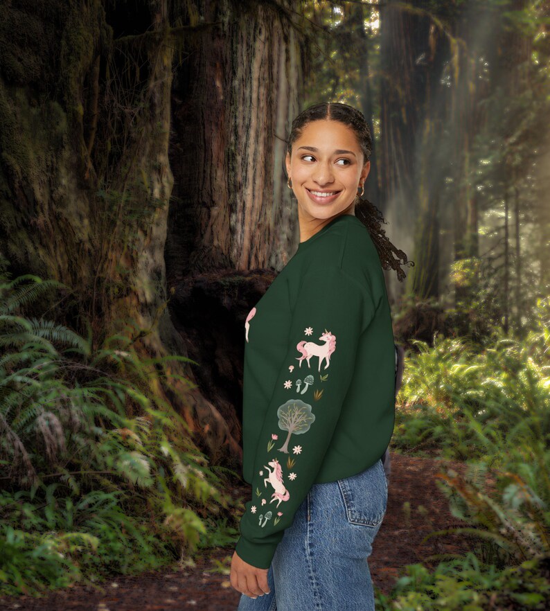May include: A dark green sweatshirt with long sleeves featuring a pink unicorn and floral design. The sweatshirt is worn by a person standing in a forest setting.