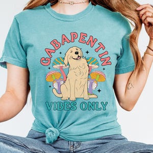 K&ouml;nnte beinhalten: Ein t&uuml;rkisfarbenes T-Shirt mit einem Aufdruck eines Golden Retriever-Hundes, der auf psychedelischen Pilzen sitzt. Der Text "Gabapentin Vibes Only" ist auf das Shirt gedruckt.