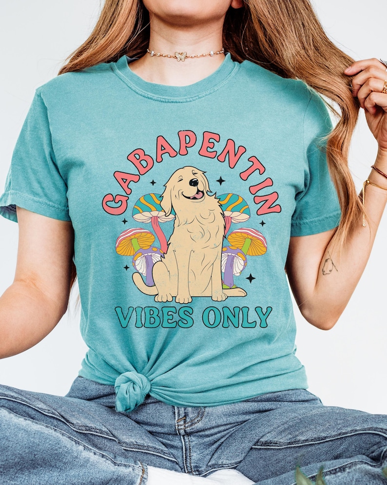 K&ouml;nnte beinhalten: Ein t&uuml;rkisfarbenes T-Shirt mit einem Aufdruck eines Golden Retriever-Hundes, der auf psychedelischen Pilzen sitzt. Der Text "Gabapentin Vibes Only" ist auf das Shirt gedruckt.