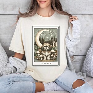 May include: A cream-colored t-shirt with a graphic of a Shih Tzu dog sitting in a forest with a crescent moon and stars above. The text "THE SHIH TZU" is printed below the image.