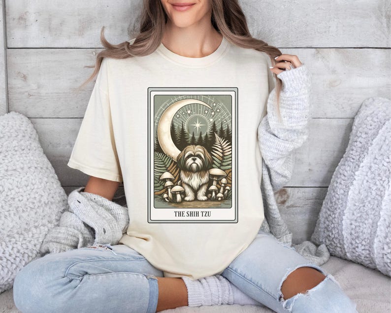 May include: A cream-colored t-shirt with a graphic of a Shih Tzu dog sitting in a forest with a crescent moon and stars above. The text "THE SHIH TZU" is printed below the image.