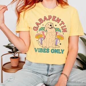 K&ouml;nnte beinhalten: Gelbes T-Shirt mit einem Grafikdruck eines Golden Retrievers, der vor Pilzen sitzt, mit dem Text "Gabapentin Vibes Only".