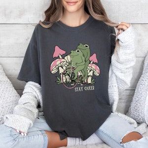 May include: A dark gray t-shirt with a graphic of two frogs riding a bicycle with pink mushrooms in the background. The text "Stay Queer" is printed below the frogs.