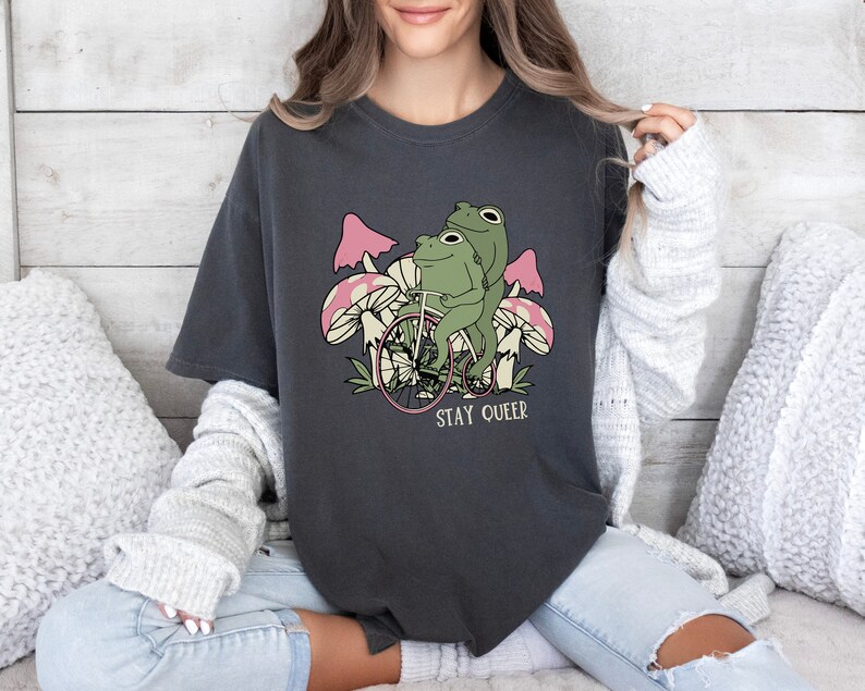 May include: A dark gray t-shirt with a graphic of two frogs riding a bicycle with pink mushrooms in the background. The text "Stay Queer" is printed below the frogs.