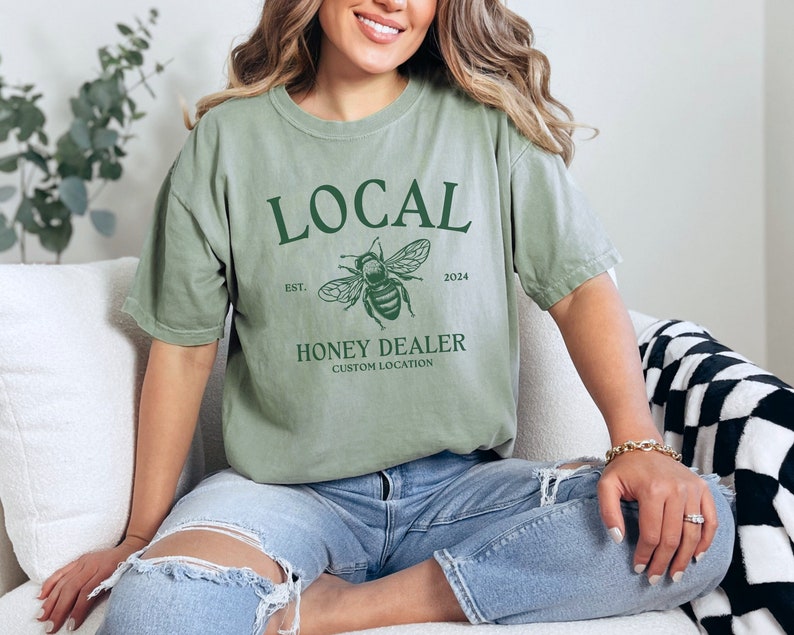 May include: A green t-shirt with the text "LOCAL" in green, a bee graphic, and the text "EST. 2024 HONEY DEALER CUSTOM LOCATION".
