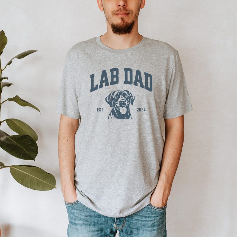 May include: A gray t-shirt with the text "Lab Dad" in blue, a black Labrador Retriever silhouette, and the text "Est 2024".