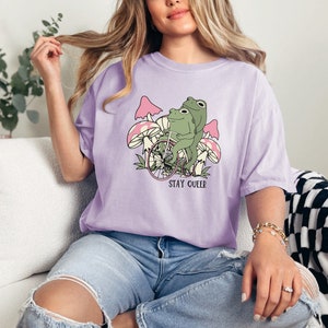 May include: A lavender t-shirt with a graphic of two frogs riding a bicycle through a field of mushrooms. The text "STAY QUEER" is printed below the image.