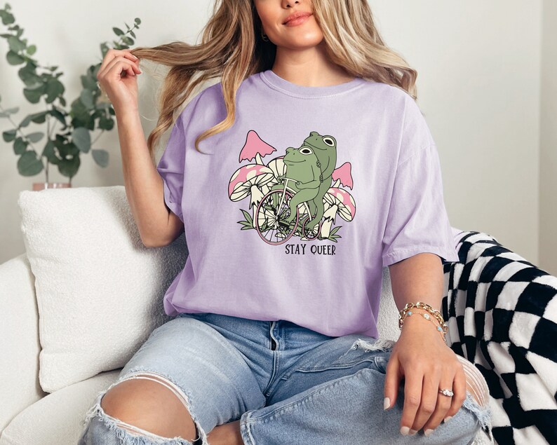 May include: A lavender t-shirt with a graphic of two frogs riding a bicycle through a field of mushrooms. The text "STAY QUEER" is printed below the image.