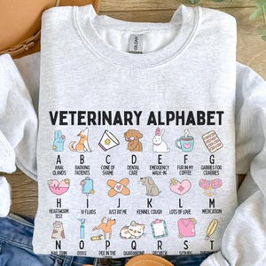 Puede incluir: Sudadera gris claro con el texto "VETERINARY ALPHABET" e ilustraciones coloridas para cada letra, incluyendo animales y símbolos médicos. La sudadera está hecha de un material suave, ideal para profesionales veterinarios.