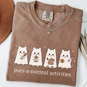 May include: A brown crewneck t-shirt featuring a graphic of four white ghost cats dressed as medical professionals. The cats hold a syringe, medicine bottle, clipboard, and thermometer with a pumpkin. The text reads purr-a-normal activities.