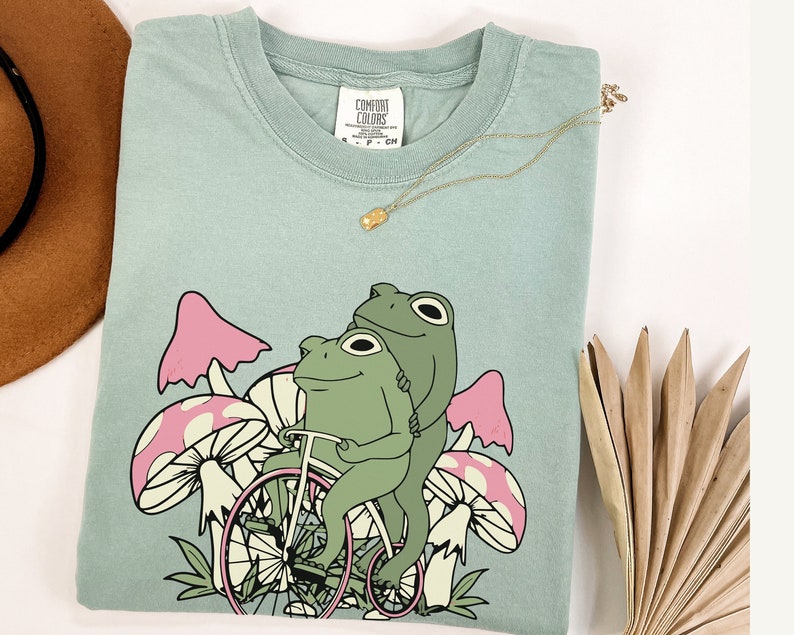 May include: A light blue t-shirt with a graphic of two green frogs riding a bicycle through a field of pink and white mushrooms. The shirt has the text "Comfort Colors" printed on the tag.