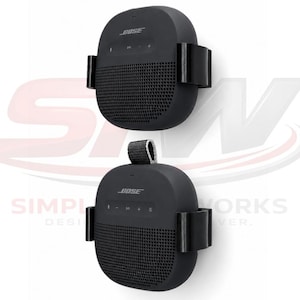 May include: Two black Bose portable speakers mounted in black plastic wall brackets. Each speaker features a rounded square shape, a perforated grille on the lower half, and control buttons on the top section. One speaker includes a small fabric loop.