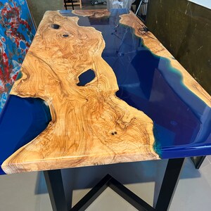 Peut inclure: Un plateau de table en bois avec une rivière en résine époxy bleue qui le traverse. La table a un piétement en métal noir.