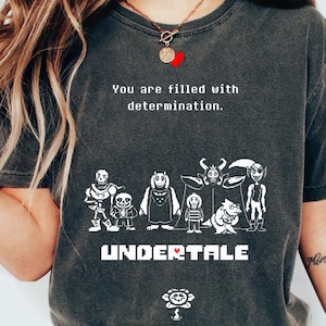 Może przedstawiać: Ciemnoszara koszulka z białym nadrukiem postaci z gry wideo Undertale. Tekst "You are filled with determination" znajduje się nad postaciami, a tytuł gry poniżej. Grafika kwiatu znajduje się na dole.