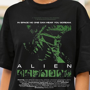 Könnte beinhalten: Ein schwarzes T-Shirt mit einem grünen Grafikdruck des Alien-Filmplakats. Das Design enthält den Text IN SPACE NO ONE CAN HEAR YOU SCREAM, den Titel ALIEN, eine Besetzungsliste und kleine quadratische Porträts der Schauspieler am unteren Rand.
