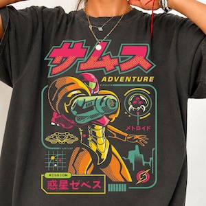 Könnte beinhalten: Ein anthrazitfarbenes T-Shirt mit einem Grafikdruck einer Figur in einem orange-gelben Anzug mit einer großen Armkanone. Das Design enthält den Text "SAMUS ADVENTURE" und japanische Schriftzeichen sowie ein kleines Kreaturensymbol und ein Raster.
