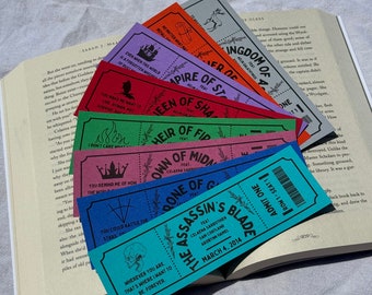 May include: A collection of colorful, ticket-shaped bookmarks with various designs and text, fanned out over an open book. The bookmarks feature phrases like "The Assassin's Blade" and "Kingdom of Ash."