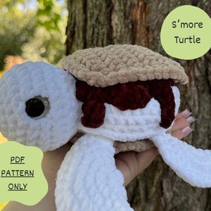 May include: A crocheted s'more turtle amigurumi toy. The turtle is white with a brown chocolate topping and a tan marshmallow shell. The toy is held in a hand and the text 'S'more Turtle' is visible in the background.