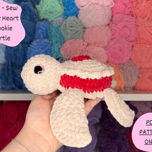 May include: A handmade, plush turtle in cream, red, and white. The turtle has a textured, soft appearance with a red stripe around the middle. The image includes text that reads "Low - Sew Jelly Heart Cookie Turtle" and "PDF Pattern Only".