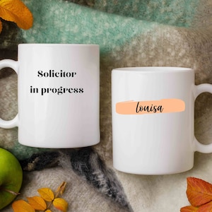 May include: Two white ceramic mugs. One mug has the text "Solicitor in progress" in black. The other mug has the name "Louisa" in black text over a peach-coloured horizontal rectangle.
