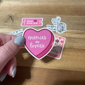 K&ouml;nnte beinhalten: Eine Sammlung von Aufklebern mit verschiedenen Designs und Texten. Der zentrale Aufkleber ist herzf&ouml;rmig und tr&auml;gt den Schriftzug "enemies to lovers" in Rosa. Weitere Aufkleber enthalten Texte wie "smut book club" und "you had me at trigger warnings."