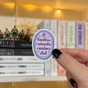 May include: An oval sticker with a light blue scalloped border and the text "hopeless romantic readers club" in a purple and white design. The sticker includes small heart and book illustrations. The sticker is held in front of a shelf of books.