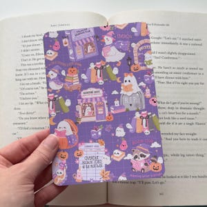May include: A rectangular bookmark with a purple grid background featuring illustrations of ghosts, ducks, raccoons, pumpkins, and books. Text includes "Haunted Reading Club," "Bookstore Ghost," "Paranormal Book Club," and "Overdue because I died at the plot twist."