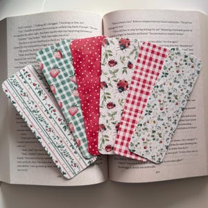 May include: A collection of six decorative bookmarks with various patterns, including strawberries, ladybugs, and gingham. The bookmarks are made of paper and are placed on an open book. The color palette includes red, green, and white.
