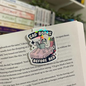 May include: A holographic bookmark with a white ghost reading a book in bed. The bookmark has the text "Sad Books Before Bed" with a rainbow glitter effect.