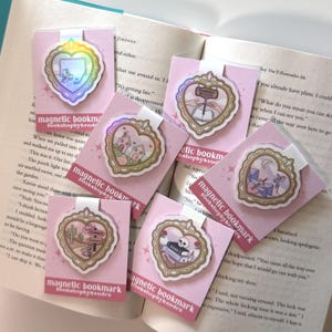 May include: A collection of magnetic bookmarks with heart-shaped designs. Each bookmark features a unique illustration within a gold-colored frame, including a computer, houses, a book, a cowboy scene, a skull, and a musical scene. The bookmarks are on pink cards with the text "magnetic bookmark bookshopbykendra".