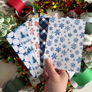 May include: A hand holds a fan of rectangular Christmas cards with various designs. The cards feature patterns of blue bows, winter scenes, gingerbread men, plaid, and blue snowflakes on a white background. Festive ribbons and tinsel surround the cards.