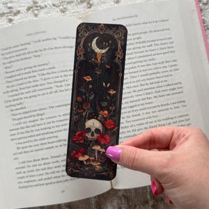 K&ouml;nnte beinhalten: Schwarzer Lesezeichen mit einem gotischen Design mit einem Totenkopf, roten Blumen, Pilzen und einem Halbmond. Das Lesezeichen wird &uuml;ber einem offenen Buch gehalten.