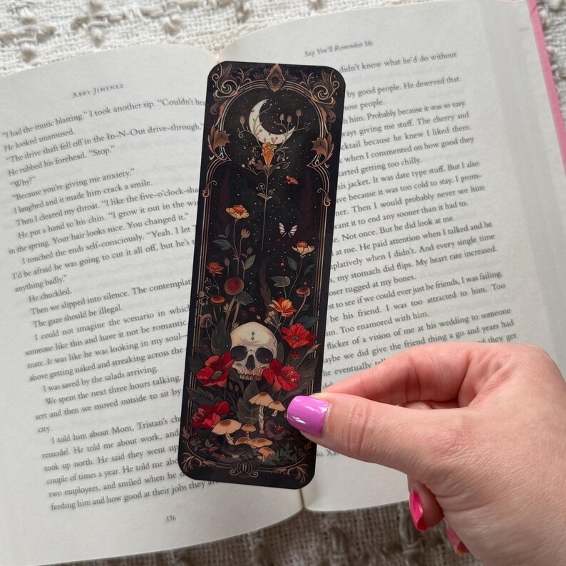 K&ouml;nnte beinhalten: Schwarzer Lesezeichen mit einem gotischen Design mit einem Totenkopf, roten Blumen, Pilzen und einem Halbmond. Das Lesezeichen wird &uuml;ber einem offenen Buch gehalten.