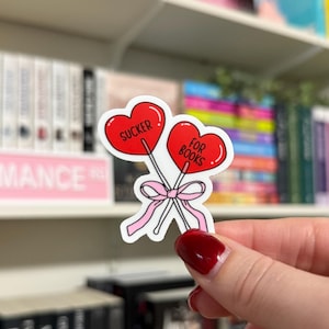 May include: Two red heart-shaped lollipops with white sticks and a pink ribbon. The lollipops have the text "SUCKER" and "FOR BOOKS" on them.