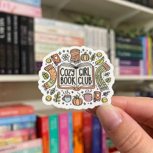 May include: A white sticker with a book, a mug, a scarf, and a pumpkin. The text on the sticker reads "Cozy Girl Book Club".
