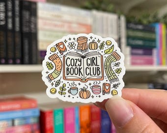 May include: A white sticker with a book, a mug, a scarf, and a pumpkin. The text on the sticker reads "Cozy Girl Book Club".