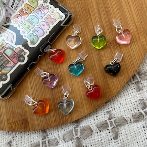 May include: Ten heart-shaped charms in various colors, including red, pink, blue, green, orange, and black. The charms are made of clear plastic and have a silver metal loop at the top. The charms are arranged on a wooden surface.