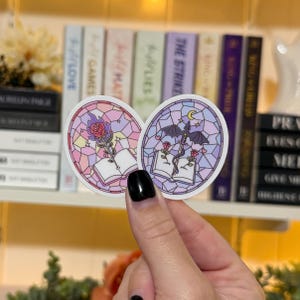 May include: Two oval stickers with stained glass designs. One features a rose with flames, the other a dragon with a sword and moon. Both have open books and roses. The stickers are in shades of pink, purple, and blue.